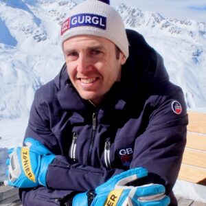 Smiling skier in a GURGL bobble hat and blue Ziener gloves leans on a wooden table with snow-capped mountains behind.