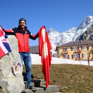 Dave 'the Rocket' Ryding unveils centenary memorial to first ever slalom race in Mürren before heading for home Man in red jacket holds a Union Jack and Swiss flag beside a stone marker, with snow-capped peaks and a hotel in the background.