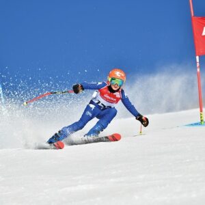 Excitement mounts as Südtirol Gardenissima slalom races return to Val Gardena Skier in a blue racing suit and red helmet carves through snow past a red "Dolomites Val Gardena" gate under a clear blue sky.