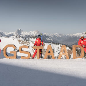 Ski race champion moves from start gates and race courses to starters and main courses in Gstaad Three skiers in red suits pose with their skis beside large wooden "#GSTAAD" letters on a snowy peak, with alpine mountains stretching behind.
