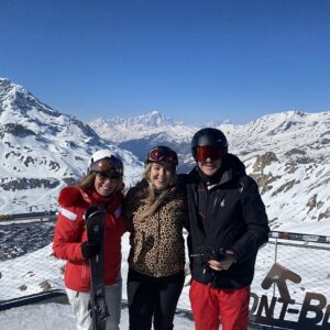 Love & Piste staff head back to the slopes of Val d’Isère Three people in ski gear pose smiling on a snowy peak with skis and snowboards, backed by snow-capped mountains and a clear blue sky.