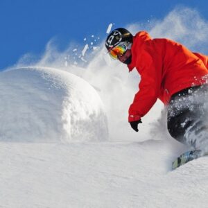 VACANCY: SnowSkool General Manager A snowboarder in a red jacket carves through deep powder, sending up a spray of snow against a clear blue sky.