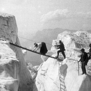Mountaineers cross a crevasse on a makeshift wooden ladder bridge over icy, snow-covered ridges in a black-and-white historical photograph.