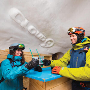 Springtime in Innsbruck - from the slopes to the sunny beer gardens Two smiling skiers in helmets and goggles clink drinks with green straws at a blue table inside a snow-walled bar.