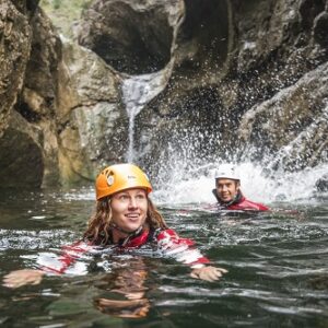 Two helmeted people in red wetsuits swim in a rocky canyon pool beneath a cascading waterfall.