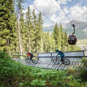 Italian ski areas set to welcome runners and cyclists for some summer fun Two mountain bikers ride across a wooden bridge through a forest while a red gondola passes overhead.