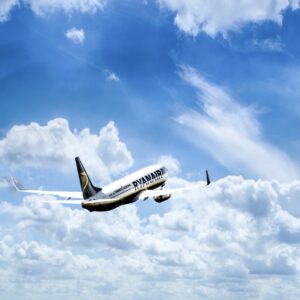 A white Ryanair passenger jet ascends through a blue sky filled with scattered white clouds.