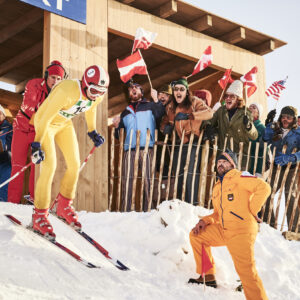 The exciting story of Franz Klammer's dramatic and historic 1976 Olympic gold medal winning run captured in a new movie A skier in yellow races past a cheering crowd waving international flags, with a man in an orange ski suit crouched in snow.