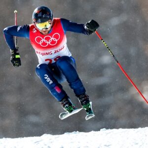 Snowsport England provides more opportunities through cross discipline development programme A skier in a GBR uniform wearing a Beijing 2022 Olympic bib and a Union Jack helmet races downhill on snow, kicking up a spray.