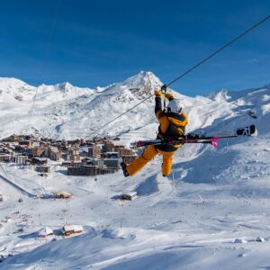 Teams challenged to compete for the 3 Valley Rally Cup as part of youth charity's 20th anniversary celebrations A skier in a yellow snowsuit rides a zip wire above a snow-covered alpine resort with clustered buildings and distant mountain peaks.