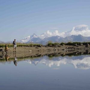 Take a voyage of discovery through all the activities on offer in Arc 1950 this summer Two people stand on a stony shore beside a still pool, with a snow-capped mountain and clouds reflected in the water.