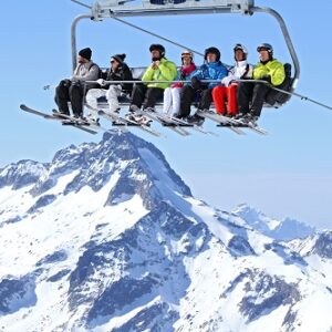 Six skiers ride a chairlift above snow-capped alpine peaks under a clear blue sky, skis dangling below their feet.