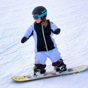 A young snowboarder in a black helmet, goggles and black and white jacket glides down a snowy slope on a yellow board.