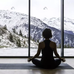 A woman sits cross-legged in a meditative pose on a yoga mat, gazing through large windows at snow-covered mountain peaks and pine forests.
