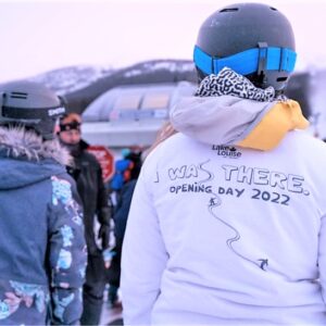A person in a white "Lake Louise" hoodie reading "I WAS THERE. OPENING DAY 2022" stands among other helmeted skiers on a snowy mountainside.