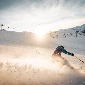 A skier in a blue jacket carves down a sunlit slope, sending powder flying, with a ski lift and mountains visible in the distance.