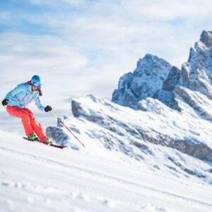 Val Gardena continues to bolster its legendary and historic attractions A skier in a light blue jacket and orange trousers carves down a snowy slope beneath jagged rocky mountain peaks.