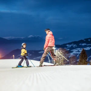 What’s new? The 22/23 ski and snowboard season in the Austrian Tirol An adult in a coral puffer jacket and a child in a yellow jacket ski across a groomed slope at dusk, with floodlit mountains in the background.