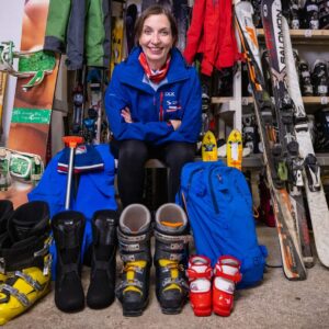 DSUK SkiBay second hand store campaign aims to raise funds to give lessons to disabled skiers and snowboarders - plus it will help save the planet A smiling woman in a blue jacket sits surrounded by ski equipment, including boots, skis, and a blue backpack arranged on the floor.