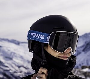 Goggles A skier in a black helmet and POW-branded goggles stands before snow-capped mountains under a grey sky.