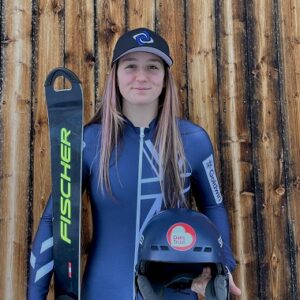 British racer teams up with Dan's Trust charity A young woman in a blue top and cap holds a Fischer ski and a helmet against a wooden slatted wall.