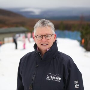 Cairngorm ski and snowboard area funicular back on track A grey-haired person in glasses wears a Cairngorm Mountain Scotland jacket on a snowy slope with distant hills and other figures in the background.