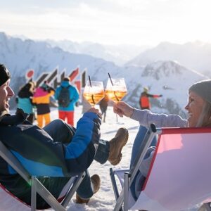Italian superstar to perform at Ischgl’s Top of the Mountain Closing Concert Two people recline in deck chairs, clinking glasses of orange cocktails against a backdrop of snow-capped mountains and skiers.