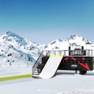 All aboard the school bus with a dry slope A converted black bus with "JIBWORX" branding sits in a snowy mountain landscape, featuring a viewing platform and a rear ramp for snowboarding tricks.