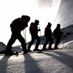 What’s new in Aosta? Silhouetted skiers with backpacks ascend a snowy slope, their long shadows cast across the snow under a bright low sun.