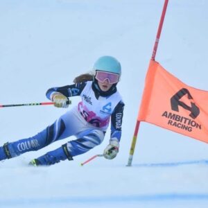 Carrick-Smith twins star in Pila A skier in a blue and white racing suit and bib carves sharply around a red gate flying an orange "Ambition Racing" flag on snow.
