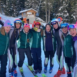 A cheerful group of skiers in green, black, and white race suits and helmets pose arm-in-arm on snow outside a wooden building marked "FLAINE.
