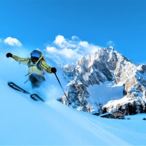 ‘Peak of Taste’ back on the Courmayeur menu A skier in a pale jacket carves through deep powder on a snowy slope, with rugged snow-capped peaks and a cluster of buildings behind under a clear blue sky.