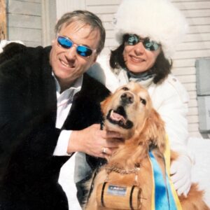 Dogs in the snow - Severin's hotel takes the lead in Lech A smiling couple in sunglasses and winter coats pose with a golden retriever wearing a small wooden barrel collar and a blue and yellow sash in the snow.