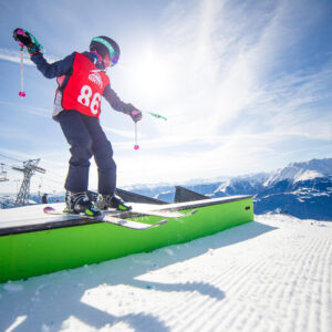 The BRITS Snowboard and Freeski championsips return to Cairngorm in April A skier in a red number 86 bib balances on a green terrain park rail against a backdrop of snow-capped mountains and ski lifts.