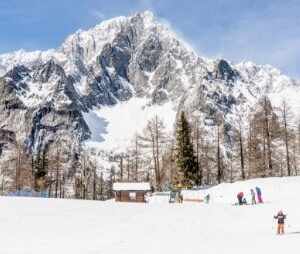 Courmayeur Mont Blanc’s hosts special Family Easter Ski Week Skiers gather on a snowy slope before a wooden hut and bare trees, with a jagged snow-capped mountain rising against a clear blue sky.