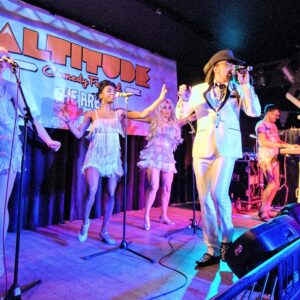 Award winning comedy festival heads to Mayrhofen Performers in fringed dresses and a suited singer wearing a hat perform on stage beneath an Altitude Comedy Festival backdrop under blue lighting.