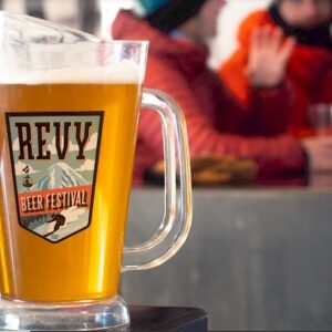 Revelstoke's beer festival invites skiers and snowboarders to celebrate end of season in style A glass jug of beer labelled "Revy Beer Festival" sits on a counter, with blurred figures in winter clothing behind it.