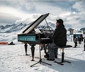 Music and film fans invited to Val d’Isère for spring festival of cultural events A man in a black coat plays a grand piano marked "AVOUS DE JOUER!" on a snowy mountainside, with onlookers and a young skier nearby.