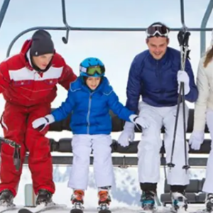 A family of four in colourful ski gear pose together on a snowy slope beneath a chairlift.