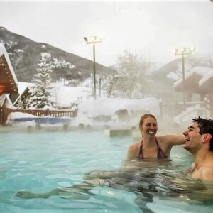 A man and woman laugh together in a steaming outdoor pool surrounded by snow-covered chalets and mountains.