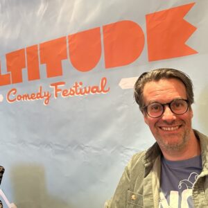 Have you heard the one about the comedy show in the snow...? A smiling man wearing glasses and a green jacket stands in front of a pale blue backdrop featuring the orange "Altifolk Comedy Festival" logo.