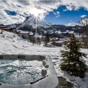 Plan ahead for 2023/24 School holiday breaks advises Oxford Ski Company A bubbling outdoor hot tub sits in the snow, looking out over snow-covered chalets and sunlit mountains.