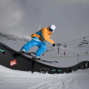 Sustainability is key to Ski Beat’s 2023/24 ski and snowboard holidays Skier in orange and blue outerwear balances on a black Snowbox rail with a ski lift and snow-covered mountains in the background.