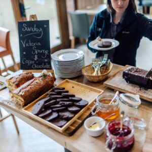 Early birds save money on 2023/24 ski and snowboard holidays with VIP SKI A server slices a chocolate cake beside a chalkboard reading "Sunday Victoria Sponge & Coffee & Chocolate," with pastries, cookies, jam and honey on the table.