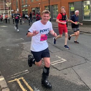 A man wearing a medical boot runs along a city street alongside other participants in a road race, smiling and waving.