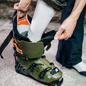 A person in a white sock slides their foot into an olive green K2 ski boot, with hands pulling open the cuff.