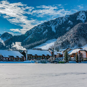 Crystal Ski Holidays announces Spain and Slovenia as new destinations for winter 2023/24 Snow-covered buildings including a hotel with a visible "HOTEL SOMMER" sign sit at the base of forested, snow-dusted mountains under a blue, partly cloudy sky.