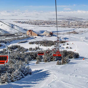 Turkish delight: a “city and ski” combo Red chairlifts descend above snow-dusted pines towards a valley town, with distant snow-capped mountains under a clear blue sky.