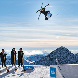 The stage is set for the world's best freeskiers on the Stubai Glacier A skier soars through an inverted aerial trick above five traditionally-dressed musicians playing long alphorns on a snowy peak.