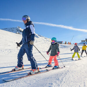 Easter Family Ski Holidays still available with Action Outdoors A skiing instructor in a navy jacket leads a line of children in colourful ski gear down a sunlit, snow-covered slope.